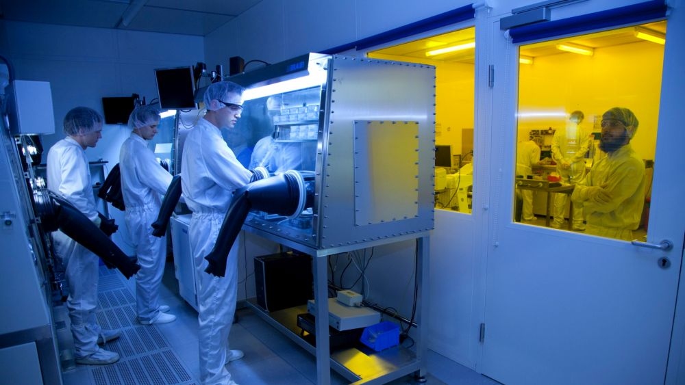 Researchers in cleanroom suits operate equipment in a lab; blue lighting, yellow room visible.
